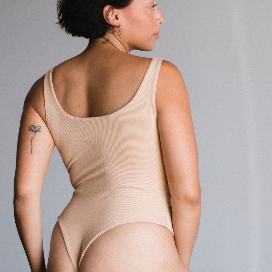 Back view of model in Ada nude, a beige tone, bodysuit. Scoop neck design. Thong style.